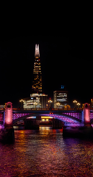 London Bridge illuminated with purple neon lights reflecting its light on the water of the River Thames and The Shard skyscraper and office buildings fully illuminated with their windows.