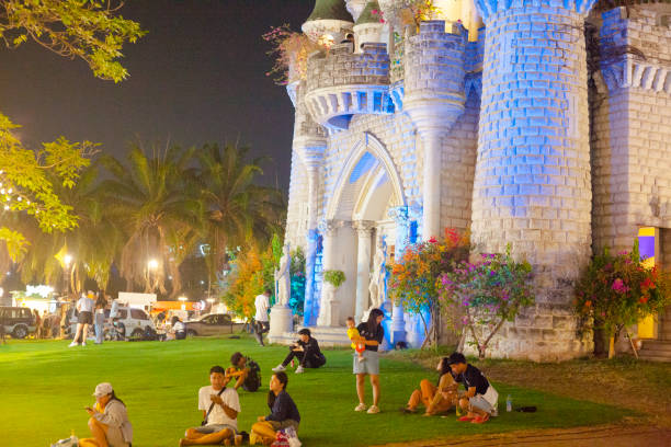 Relaxing thai people in grass in front of miniature castle on  night market Danneramit in Bangkok.  Castle is made in euopean style and was once a club house and now exhibition house at night and train market. People are standing or sitting in groups on  grass. Castle has some reflections of blue neon lights at wall. One woman is holding a child on arm. Castle is one of attractions of night market