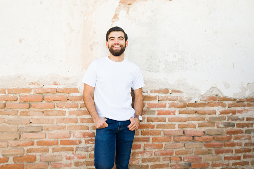 Cheerful young hispanic man wearing a white t-shirt mockup stands confidently in front of a rustic brick wall, exuding casual charm