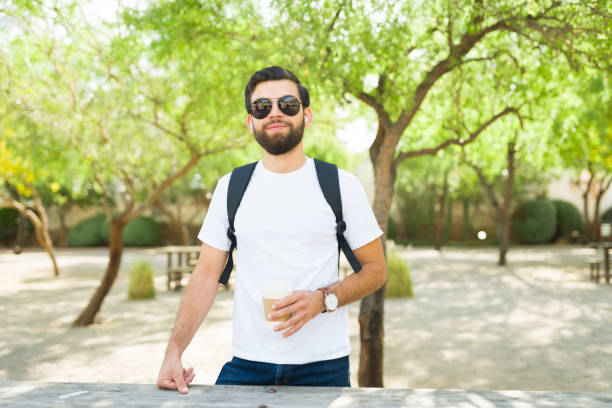 Young hispanic man exuding confidence with a backpack, wearing a white t-shirt for mockup, sunglasses, and smiling in a sunny park setting