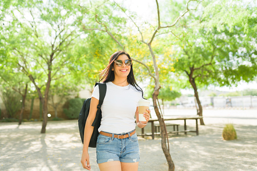 Smiling young hispanic woman wearing a white t-shirt and sunglasses, strolling in a sunny park with coffee and a backpack