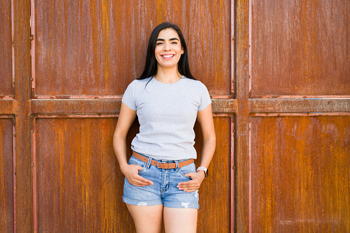 Cheerful young latin woman posing in front of a metallic door, sporting a plain grey t-shirt perfect for brand mockups