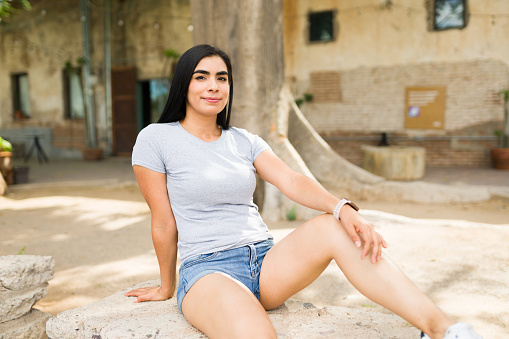 Confident young latin woman wearing a casual gray t-shirt and denim shorts, sitting on a stone bench in a sunny park setting
