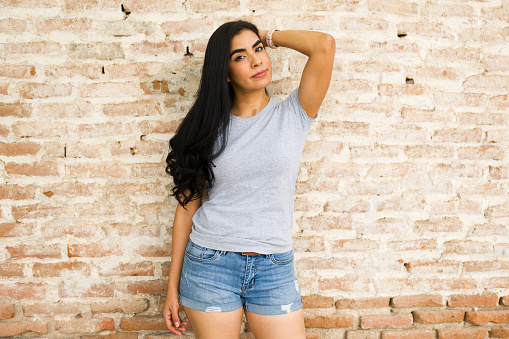 Stylish young latin woman models a plain gray t-shirt perfect for mockup designs, standing confidently with a brick wall backdrop