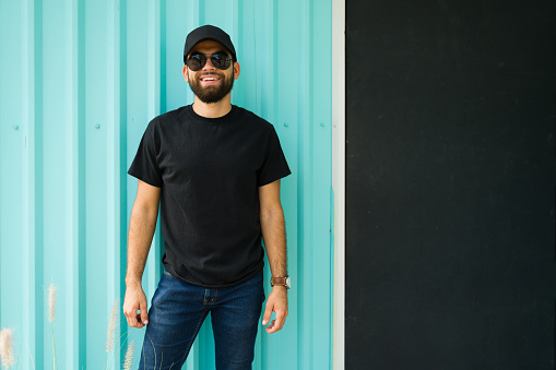 Confident latin man in a casual black t-shirt and sunglasses standing against a lively turquoise wall with a cheerful attitude