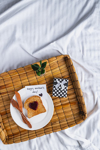 A rustic basket tray with happy mother's day greeting card, a little gift in a box, and a toast of bread with heart shape made out of jam, all over a bed on wrinkled sheet with space for text and copy