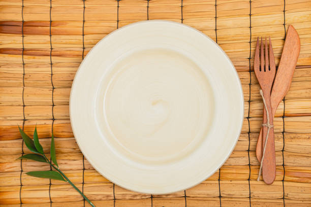 Flatlay lay of round plate mockup over a rustic basket texture with a green twig and a pair of wooden fork and knife
