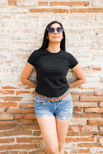Stylish hispanic woman wearing a plain black t-shirt and denim shorts poses confidently against a rustic brick wall background