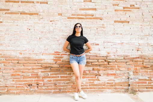 Attractive latin woman in black t-shirt and denim shorts, striking a full length pose with sunglasses in front of a textured brick wall, ideal for mockup designs