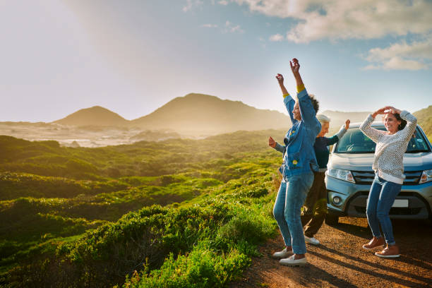 mature women cheering by their car parked by a scenic coast during a road trip - aankomst-fotos stockfoto's en -beelden