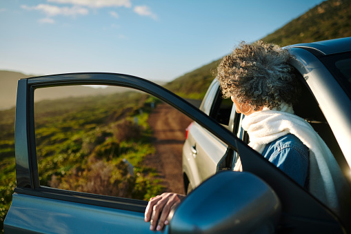Mature woman getting out of a car on a road through scenic countryside during a road trip with friends in summer