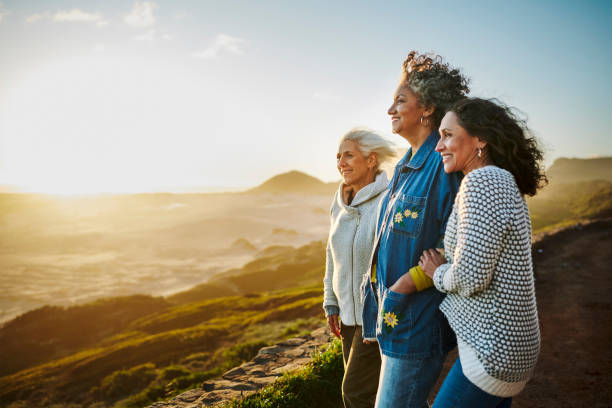 smiling mature friends standing arm in arm by a scenic coast during a road trip - mulheres-maduras imagens e fotografias de stock