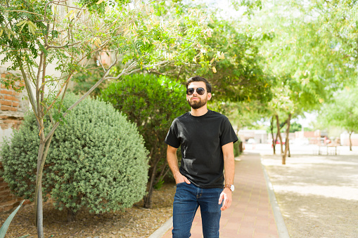 Portrait of a young hispanic man wearing sunglasses and a black t-shirt, posing for a mockup against a natural park backdrop with a relaxed stance