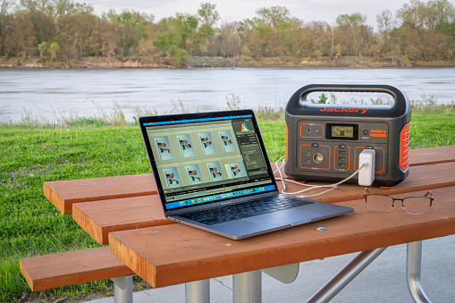 Brownville, NE, USA - April 22, 2024: Editing pictures on MacBook Pro laptop connected to Jackery power station, picnic table on a shore of the Missouri River.
