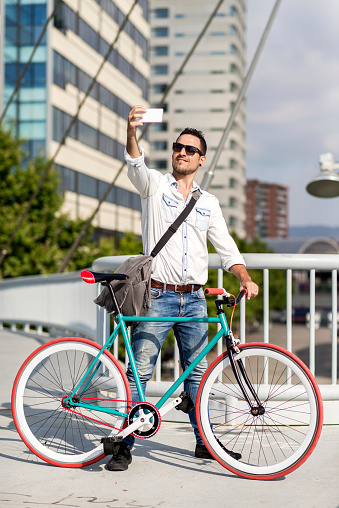 Confident man in a casual outfit takes a selfie with a smartphone while leaning on his fixed-gear bike on an urban bridge.
