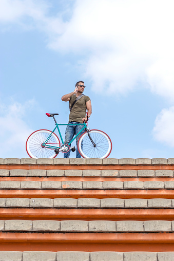 Confident man on rooftop holding bicycle while talking on the phone, against a clear sky, suggesting an urban lifestyle and connectivity