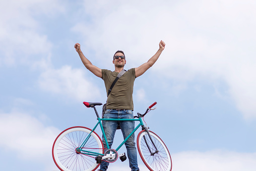 Exuberant man in casual wear lifts arms high in victory, standing with a stylish bicycle under open sky, exuding freedom and success