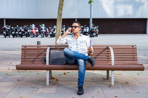 Stylish man in casual attire speaking on the phone while sitting on a wooden bench, with motorcycles in the background