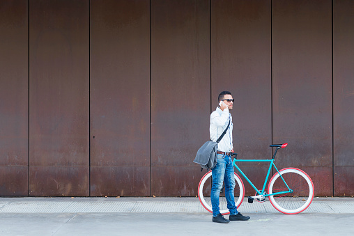 Stylish man with sunglasses and a messenger bag standing by a red-wheeled bike, making a phone call against a rusted metal wall