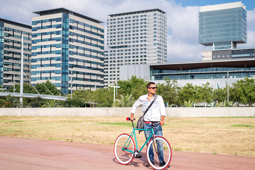 A stylish man stands with his red and turquoise bicycle in an urban park with modern buildings in the background, exuding urban lifestyle vibes