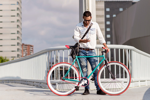 Fashionable man in white shirt and sunglasses stands with a colorful bicycle on an urban bridge, high-rise buildings in the background
