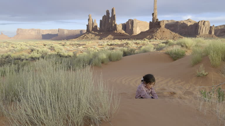 Two Year Old Navajo Boy with Traditional Hair and Pony Tail Tied with White Yarn Playing in Sand in Front of Totem Pole Rock Formation at Monument Valley Tribal Park Under Dramatic Sunset Skies
