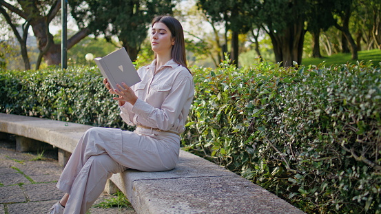 Relaxed girl looking book in beautiful green park eating apple. Calm female reader enjoy reading hobby surrounded by vivid foliage. Young woman studying novel in quiet outdoor environment at weekend.