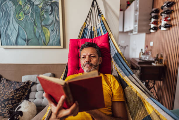istock Mature man reading a book lying down on hammock at home 2150336893