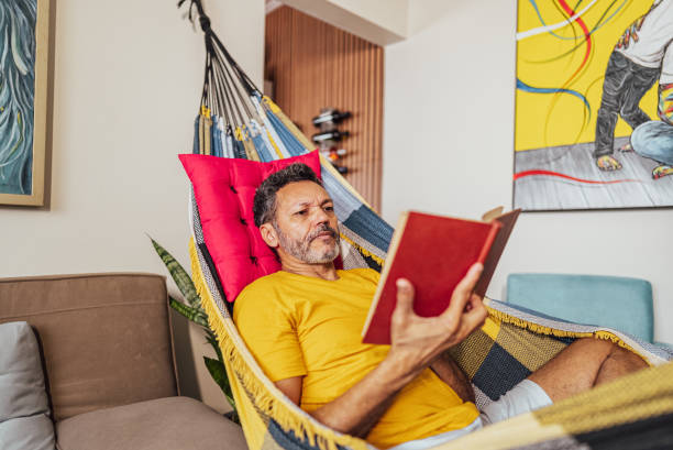 istock Mature man reading a book lying down on hammock at home 2150338463