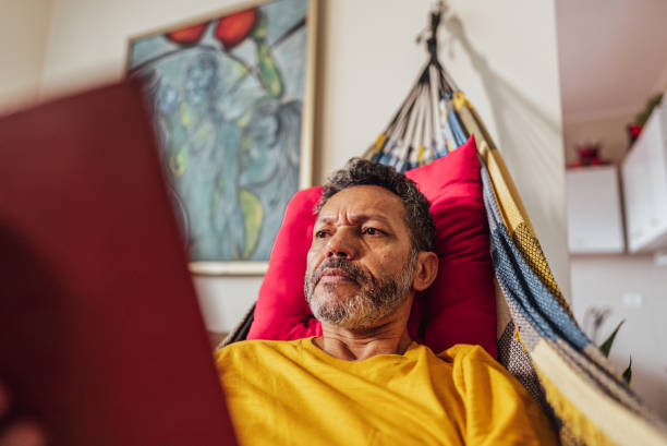 istock Mature man reading a book lying down on hammock at home 2150338464