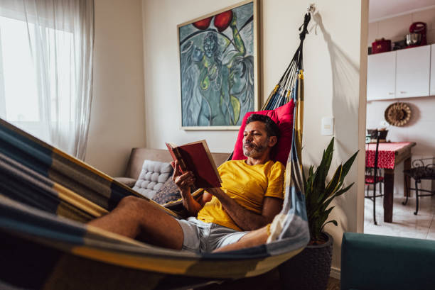 istock Mature man reading a book lying down on hammock at home 2150338465