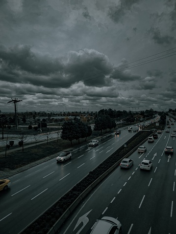 A snapshot of life’s perpetual motion, this photograph captures the essence of daily hustle as vehicles traverse the highway under a brooding sky. The overcast heavens, painted with shades of grey, loom over the earth, hinting at the imminent downpour. Despite the threat of a storm, the highway remains a vein of life, pulsing with the steady flow of cars—each a capsule of stories on their journey. The verdant flanks of the road stand in contrast, a reminder of nature’s calm amidst human urgency. This scene is a powerful portrayal of resilience and the undeterred spirit of movement, no matter the weather’s whims.