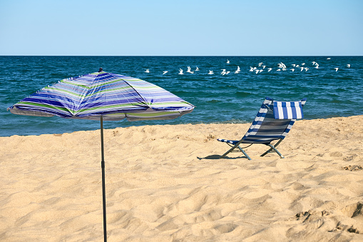 Relaxing beach scene with a blue and white striped beach chair and a blue and green striped umbrella under a clear blue sky with white clouds and a flock of seagulls flying overhead.