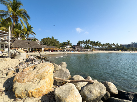 La Cruz de Huanacaxtle, Nayarit, Mexico - 2024-04-08: On a sun-drenched day at Playa la Manzanilla, tranquil waters lap against a rocky shore, while palm trees and thatched umbrellas offer a picturesque tropical backdrop for beachgoers enjoying the coastal paradise.