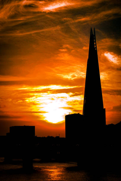 View of the silhouette of the Shard in London during sunrise. The colour of the sky and the clouds gives it a warm summer feeling. Thames river seen on the foreground