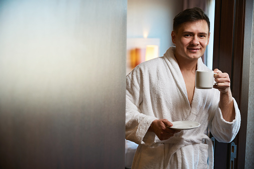 Smiling adult male in bathrobe holding ceramic cup and saucer in hands while leaning against door frame