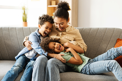 Funny African American children enjoying, fooling around with their mother, sitting on sofa at home. Concept of childhood, motherhood, family, fun
