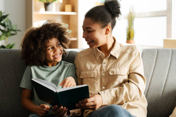 istock Smiling young mother sitting next to her little daughter on sofa at home while reading book 2150361579