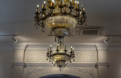 Bangkok, Thailand - Apr 15, 2024 - Glittering crystal chandeliers decorated hanging on the ceiling with Arabic alphabet decorative on wall background. Interior design and room decoration with Antique chandeliers in Bang O Mosque, Space for text, Selective focus.