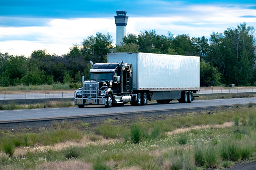 A contemporary take on a classic semi-truck design. This black 18-wheeler is pulling a plain white dry box trailer on a divided highway. You can see an air traffic control tower in the background.