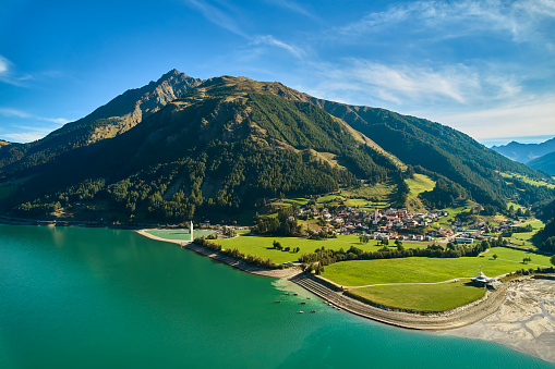 Village on the mountain near the lake (Reschensee). Kite school on the shore and church tower in the water. Aerial view at sunny noon. Italy, Vinschgau, Graun.