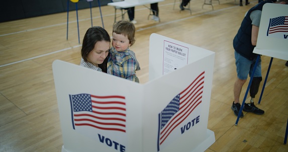 Female voter with baby on hands stands at voting booth. American citizens come to vote in polling station. Political races of US presidential candidates. National Election Day. Civic duty. Dolly shot.