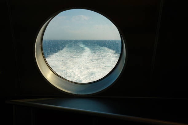 Scenic view through round porthole on cruise ship, looking out at sea and distant coastal city on sunny day