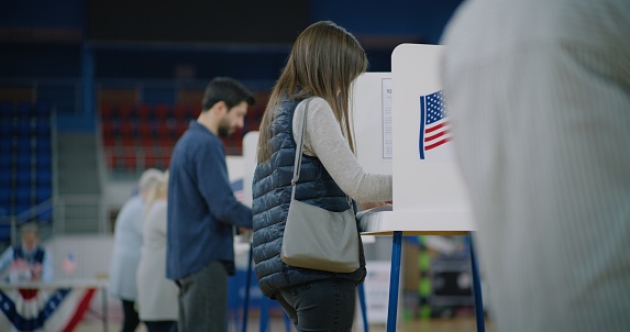 Caucasian female voter comes with bulletin to voting booth. American citizens come to vote in polling station. Political races of US presidential candidates. National Election Day in United States.