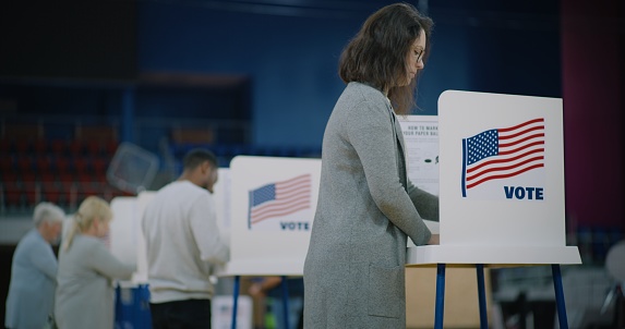 Caucasian female voter comes with bulletin to voting booth. American citizens come to vote in polling station. Political races of US presidential candidates. National Election Day in United States.