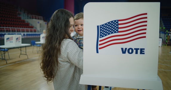 Woman with child on hands comes to voting booth. American citizens come to vote in polling station. Political races of US presidential candidates. National Election Day in United States. Dolly shot.