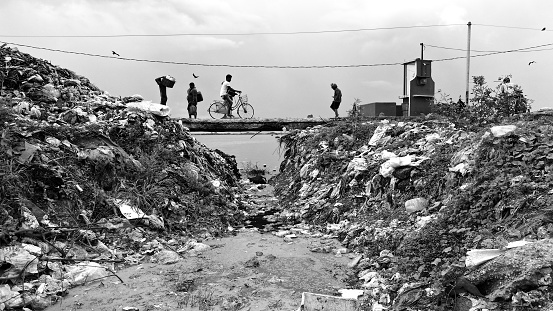 This compelling black and white photograph captures a poignant moment of daily life framed by the stark reality of environmental degradation. A small group of individuals traverses a makeshift path flanked by towering mounds of urban waste. The presence of birds and the person riding a bicycle adds a layer of normalcy against the backdrop of the neglected and polluted landscape, offering a powerful commentary on resilience and the human condition in the face of adversity.