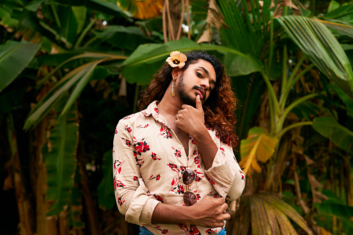 His floral shirt blends with the rich plants, expressing identity and soft masculinity. Sri Lankan gay man with a flower in his hair stands in a tropical setting, touching his chin thoughtfully.