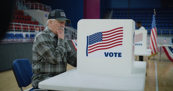 Elderly male voter sits at voting booth. American citizens come to vote in polling station. Political races of US presidential candidates. National Election Day. Civic duty and patriotism. Dolly shot.