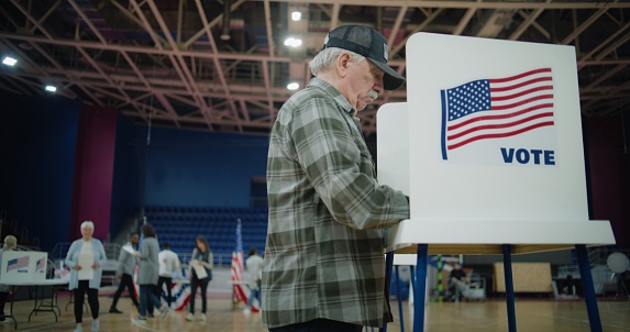 Elderly voter with bulletin in hands comes to voting booth. Diverse American citizens come to vote in polling station. Political races of US presidential candidates. National Election Day. Dolly shot.